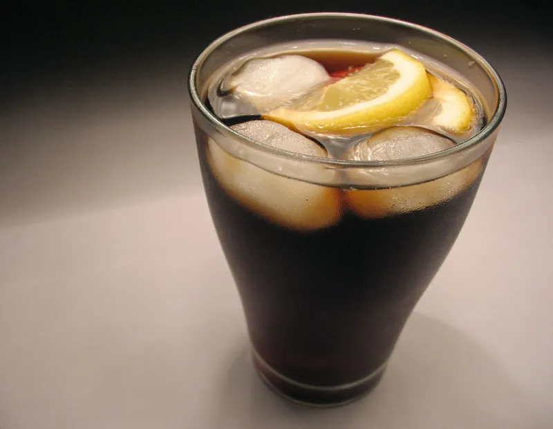 A glass of dark fizzy drink with ice cubes and a slice of lemon.
